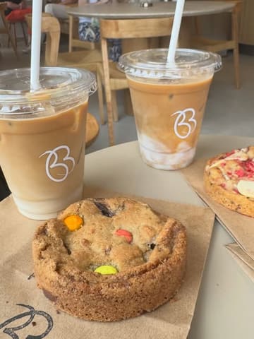 A simple, overhead flat-lay shot featuring two iced lattes and a Smarties-topped cookie on a paper bag, capturing a casual coffee shop visit.