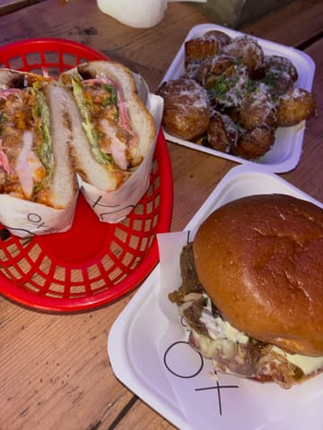 A high-angle, casual snapshot of three street food items—a sandwich, a burger, and a side of potatoes—served in paper-lined baskets on a wooden table. The image is lit with a harsh, direct flash, typical of a casual night out.
