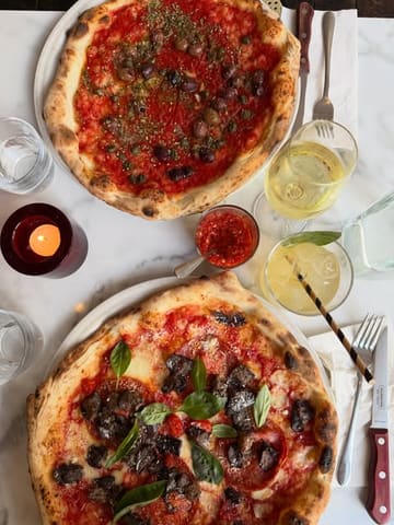 A high-quality, overhead flat-lay shot of two pizzas served on a marble table, accompanied by drinks and a candle, capturing a classic casual dining experience.