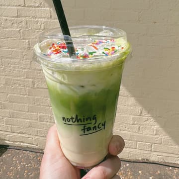 A bright, eye-level shot of a matcha latte in a clear takeaway cup, held by a hand against a neutral brick wall. The drink is topped with whipped cream and rainbow sprinkles, creating a vibrant and inviting aesthetic.