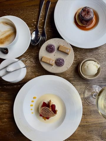 A high-quality, overhead flat-lay photograph of a multi-course lunch spread at a restaurant. The image features various plates including a cheese souffle, a dessert with poached plum, a small plate of petit fours, and a cup of coffee, all arranged neatly on a wooden table.