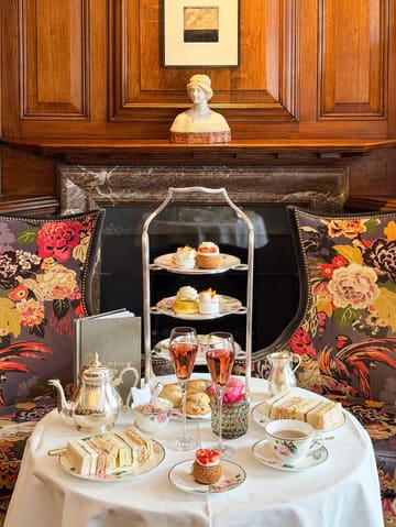 A beautifully composed, symmetrical shot of a luxury afternoon tea service set against a classic, wood-paneled fireplace. The image captures the elegance of the venue, with the tea stand, champagne flutes, and delicate pastries arranged perfectly to evoke a sense of high-end hospitality.