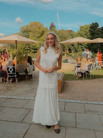 A high-quality portrait of a woman holding a glass of champagne at an outdoor event held at The Hurlingham Club. The image captures a sophisticated, social atmosphere with a shallow depth of field that keeps the focus on the subject while providing context of the event in the background.