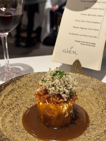 A close-up shot of a fine-dining pastry dish, likely a choux bun, served on a textured plate with a rich sauce. A menu card and a glass of red wine are visible in the background.