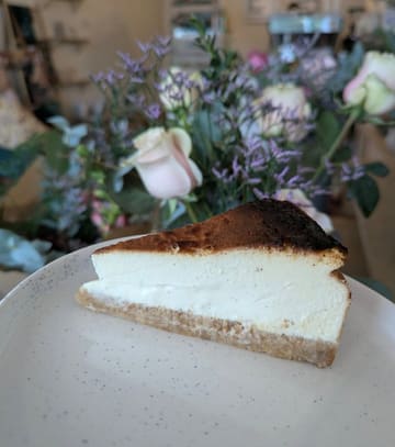 A high-quality, close-up photograph of a slice of Basque cheesecake on a speckled plate, featuring a soft-focus floral background that creates an inviting, cafe-style atmosphere.