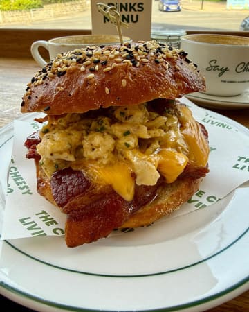 A high-quality, close-up shot of a breakfast bagel served on branded paper, likely in a cafe setting. The image effectively captures the textures of the scrambled eggs, melted cheese, and bacon, with coffee cups visible in the background to establish a relaxed breakfast atmosphere.