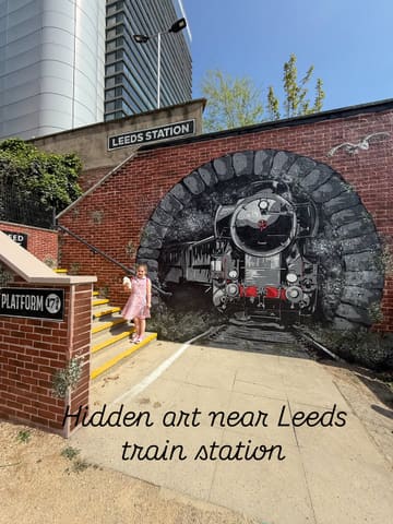 A bright, clear, and well-composed photograph featuring a young girl standing in front of a large, detailed steam train mural near Leeds train station. The natural lighting and clean composition make for an engaging travel-style image.