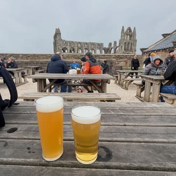 A casual, eye-level shot of two glasses of beer on a weathered wooden table at an outdoor venue, with the ruins of Whitby Abbey visible in the background.