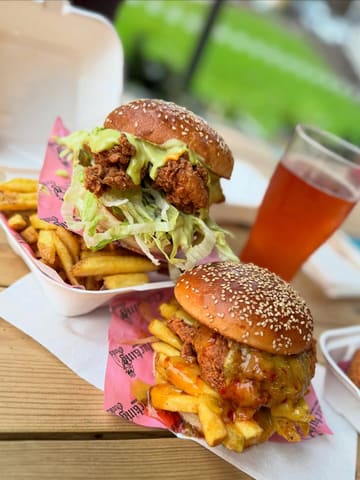 A highly appetizing, close-up shot of two fried chicken burgers served in takeaway containers on a wooden table. The natural lighting effectively highlights the crispy texture of the chicken, the vibrant greens of the lettuce, and the glossy sauces. The shallow depth of field draws the viewer's eye directly to the food, creating an indulgent and crave-worthy visual.