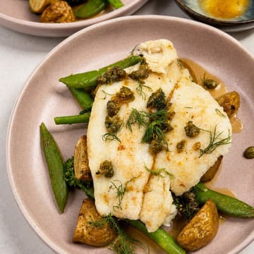 A high-quality, well-composed food photograph featuring a pan-seared lemon sole fillet served with snap peas, baby potatoes, and a burnt butter caper sauce. The lighting is bright and even, highlighting the texture of the fish and the freshness of the vegetables.