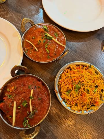 A top-down, overhead shot of two Indian curries and a bowl of pilau rice served in traditional metal dishes on a wooden table.