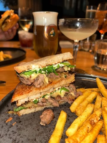 A close-up shot of a steak sandwich and fries served in a dimly lit restaurant setting, accompanied by various drinks in the background.