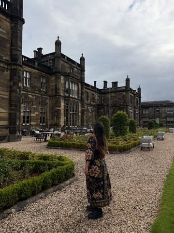 A travel-focused shot featuring a guest standing in the courtyard of Mar Hall Resort in Scotland. The image captures the historic architecture of the venue against an overcast sky, highlighting the resort's grounds and outdoor seating area.