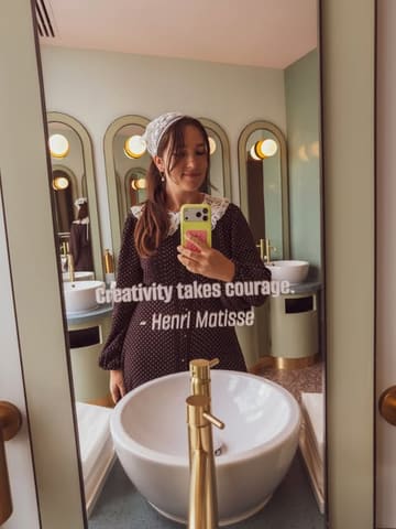 A mirror selfie taken in the stylish, modern bathroom of the Art'otel Battersea, featuring a woman in a patterned dress and headscarf. The image uses the mirror's reflection to frame the subject, highlighting the venue's interior design.