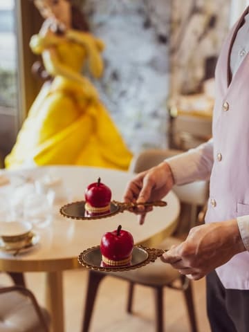A high-quality, thematic shot showcasing the 'Enchanted Apple' dessert from the Disney-themed afternoon tea at the London Hilton on Park Lane. The image uses a shallow depth of field to focus on the glossy, well-presented dessert held on a mirror, with a subtle nod to the theme in the background.