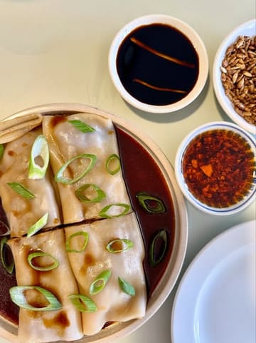 A clean, overhead shot of a dim sum dish, specifically beef cheung fun, accompanied by small bowls of chili oil and crispy toppings, presented on a light-colored table.