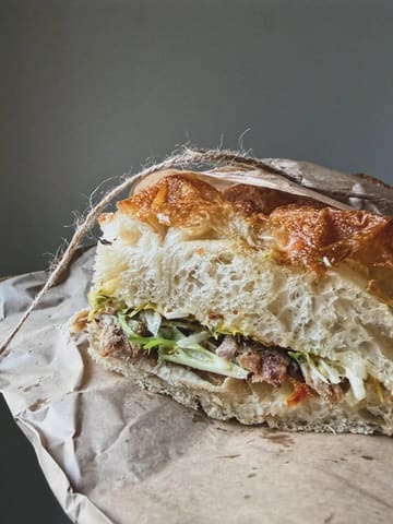 A high-quality, close-up food photograph of a pork sandwich from Lannan bakery. The image effectively captures the rustic texture of the crusty bread and the filling, presented in simple paper packaging, creating an inviting and appetizing visual.