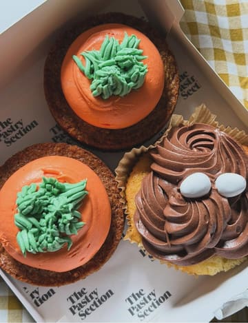 A high-quality, top-down photograph of three cupcakes presented in a branded bakery box. The image features two carrot-themed cupcakes with orange frosting and green piping, alongside a chocolate-frosted cupcake topped with small white candy eggs, highlighting a seasonal Easter aesthetic.