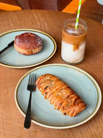 A clean, top-down shot of a cafe table featuring a spinach and feta lattice roll, a dulce de leche doughnut, and an iced coffee, showcasing the bakery's offerings.