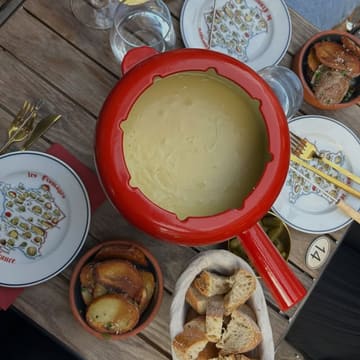 An overhead, flat-lay shot of a cheese fondue set on a wooden table, accompanied by a basket of bread and a bowl of roasted potatoes, captured in an outdoor setting.