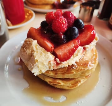 A close-up, angled shot of a stack of pancakes topped with vanilla cream, fresh strawberries, raspberries, and blueberries, drizzled with maple syrup at The Breakfast Club.