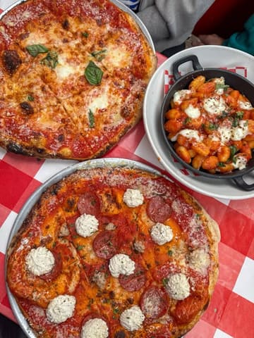 An overhead shot of a casual Italian meal featuring two pizzas and a skillet of gnocchi served on a red and white checkered tablecloth, capturing a relaxed dining atmosphere.