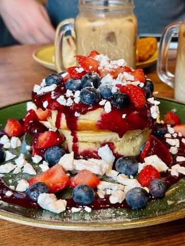 A high-quality, close-up shot of a stack of thick, fluffy pancakes topped with fresh strawberries, blueberries, crumbled meringue, and a generous drizzle of strawberry coulis, served at a brunch venue.