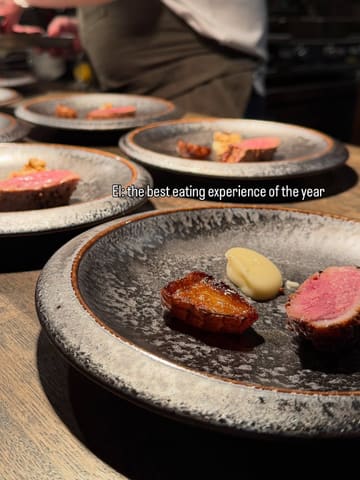 A high-quality, close-up shot of a fine-dining dish featuring a piece of seared meat, a roasted potato, and a dollop of puree, set against a blurred background of other plates.
