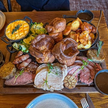 A high-quality, top-down shot of a substantial Sunday roast sharing board, featuring a trio of meats including sirloin beef, leg of lamb, and turkey, accompanied by Yorkshire puddings, roast potatoes, pigs in blankets, stuffing, and sides served in cast iron dishes. The lighting is natural and even, effectively highlighting the textures of the food.