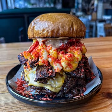 A high-quality, close-up shot of a 'West Texas Degenerate' burger, featuring a quadruple beef patty, American cheese, slow-cooked BBQ brisket, mac and cheese, and flaming hot Cheetos dust. The image is well-lit and sharply focused, highlighting the textures of the ingredients.