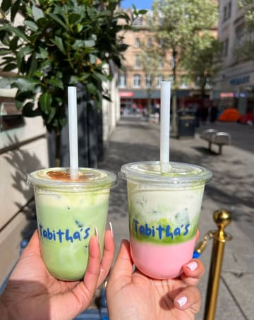 A bright, outdoor shot featuring two layered matcha lattes held up against a street background, showcasing the vibrant green and pink colours of the drinks.
