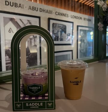 A close-up shot of two iced drinks from Saddle London, featuring an iced matcha latte in a branded arch-shaped holder and an iced Spanish latte, placed on a counter with a branded wall in the background.