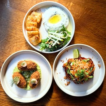 A high-quality, overhead shot of three plates of food on a wooden table, likely from a restaurant review. The lighting is natural and flattering, highlighting the textures of the dishes, which include halloumi, chicken satay, and croquettes. The composition is balanced and clean, making the food look appealing and well-presented.