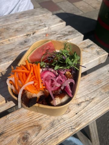 A top-down shot of a BBQ pork belly rice bowl served in a cardboard takeaway container, placed on a wooden outdoor table. The dish features pork belly, pickled carrots, pickled red onions, a slice of watermelon, and microgreens.