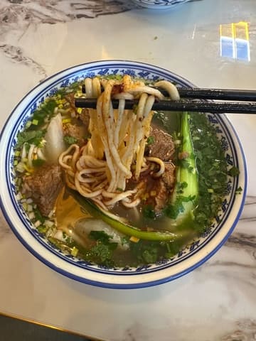 A close-up, top-down shot of a bowl of beef noodle soup being lifted with chopsticks, showcasing the texture of the noodles and the clarity of the broth.