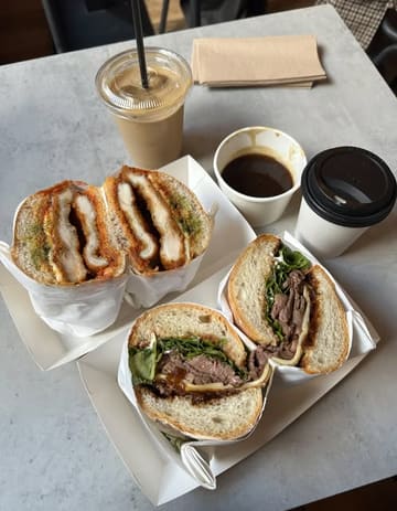 A high-quality, overhead food photography shot featuring two large, filled sandwiches (a French Dip and a Chicken Parm) alongside a coffee and a takeaway cup, likely gravy. The image is clean, well-lit, and focuses on the textures of the food.