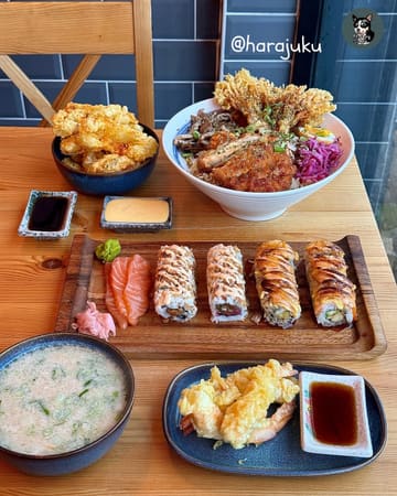 A flat-lay photograph of a Japanese meal spread, featuring sushi rolls, a large rice bowl, tempura shrimp, and a bowl of soup on a wooden table.