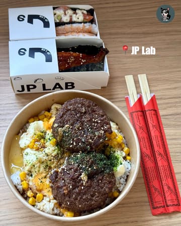 A top-down, well-lit photograph showcasing a takeaway meal from JP Lab. The image features a selection of sushi tacos in branded boxes alongside a hearty bowl of wagyu beef patty rice topped with egg. The composition is clean and focused, effectively highlighting the textures of the food.