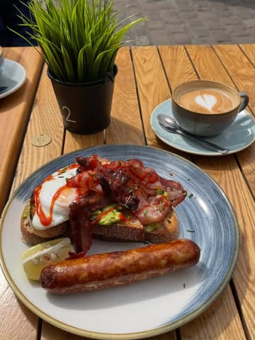A casual, natural-light shot of a brunch plate featuring avocado on sourdough, bacon, sausage, and a fried egg, accompanied by a flat white, set on an outdoor wooden table.