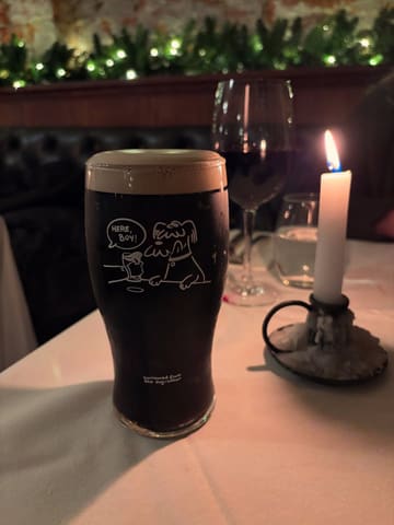 A moody, atmospheric shot of a pint of Guinness served in a branded glass, set against a candlelit table in a pub environment. The image captures a cozy, intimate vibe with a shallow depth of field that highlights the glass while blurring the background, effectively showcasing the venue's attention to detail.