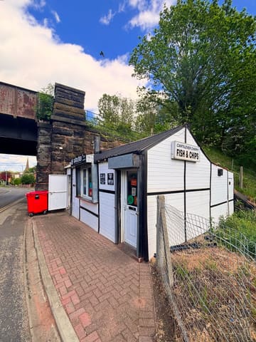 A clear, eye-level exterior shot of 'The Cantilever Chippy,' a unique, shed-like fish and chip shop situated beneath a railway bridge. The image effectively communicates the venue's compact size and historic, quirky character, providing a strong sense of place.