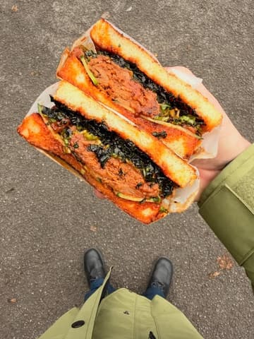 A first-person perspective shot of a char siu pork belly sandwich on Japanese milk bread, held in hand against a pavement background. The image effectively showcases the texture and filling of the sandwich.