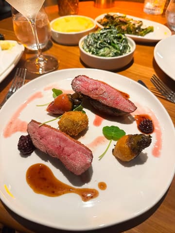 A high-angle, close-up shot of a restaurant table featuring a main course of duck breast with a croquette, accompanied by side dishes including mashed potato, creamy spinach, and tender stem broccoli. The image captures the ambient, warm atmosphere of the venue, though the lighting is heavily skewed towards orange tones.