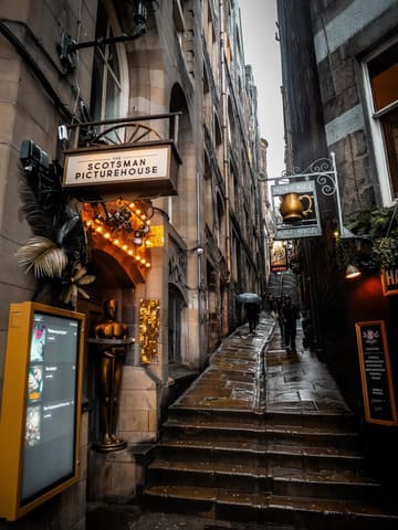 A moody, atmospheric, and high-quality photograph of Fleshmarket Close in Edinburgh, capturing the historic, narrow alleyway with signage for The Scotsman Picturehouse and The Halfway House. The image effectively conveys the unique, dark, and historic charm of the location.