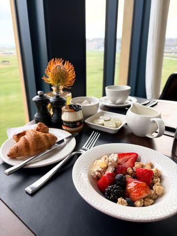 A high-quality, well-composed shot of a hotel breakfast spread featuring a croissant, a bowl of fruit and granola, and various condiments, set against a scenic window view of a golf course.