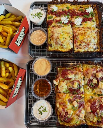 A high-quality, overhead flat-lay photograph of two Detroit-style pizzas and two portions of fries, presented on metal wire racks and branded takeaway boxes. The lighting is bright and even, effectively highlighting the textures of the crispy crusts, melted cheese, and various toppings.