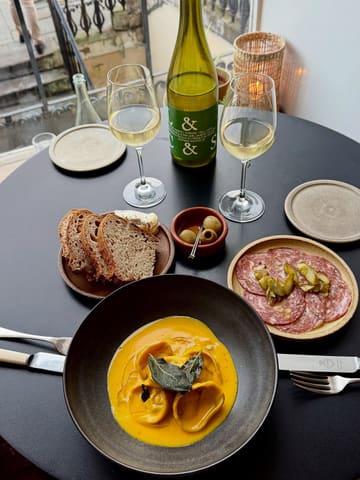 A high-quality, overhead flat-lay shot of a meal at Spry Wines in Edinburgh, featuring a bowl of pasta, charcuterie, bread, olives, and white wine. The lighting is natural and soft, highlighting the textures of the food and the inviting atmosphere of the venue.