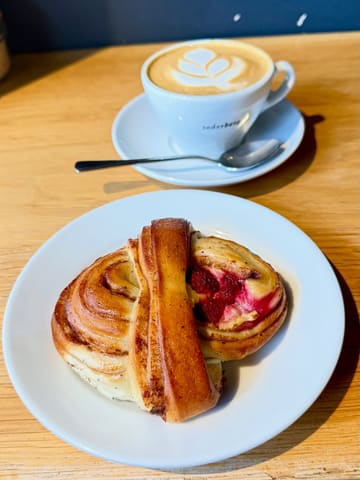 A close-up, high-angle shot of a Swedish-style pastry with a raspberry filling, accompanied by a latte with latte art, set on a wooden table at a bakery.