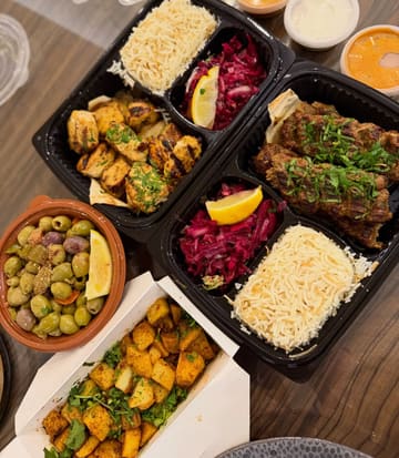 A top-down, flat-lay photograph of a Lebanese takeaway spread, featuring containers of lamb kafta, shish taouk, rice, salad, olives, and batata harra on a wooden table.