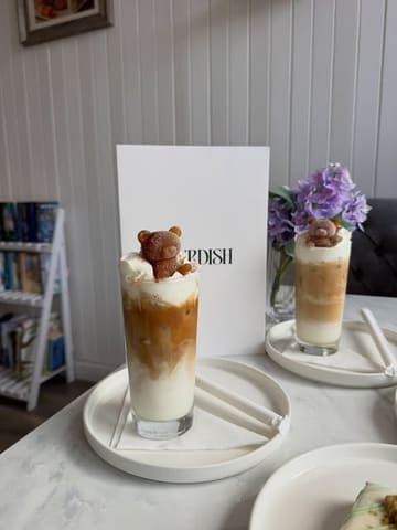 A bright, well-composed shot of two iced lattes featuring cute teddy bear-shaped ice cubes, set in a cozy cafe environment.
